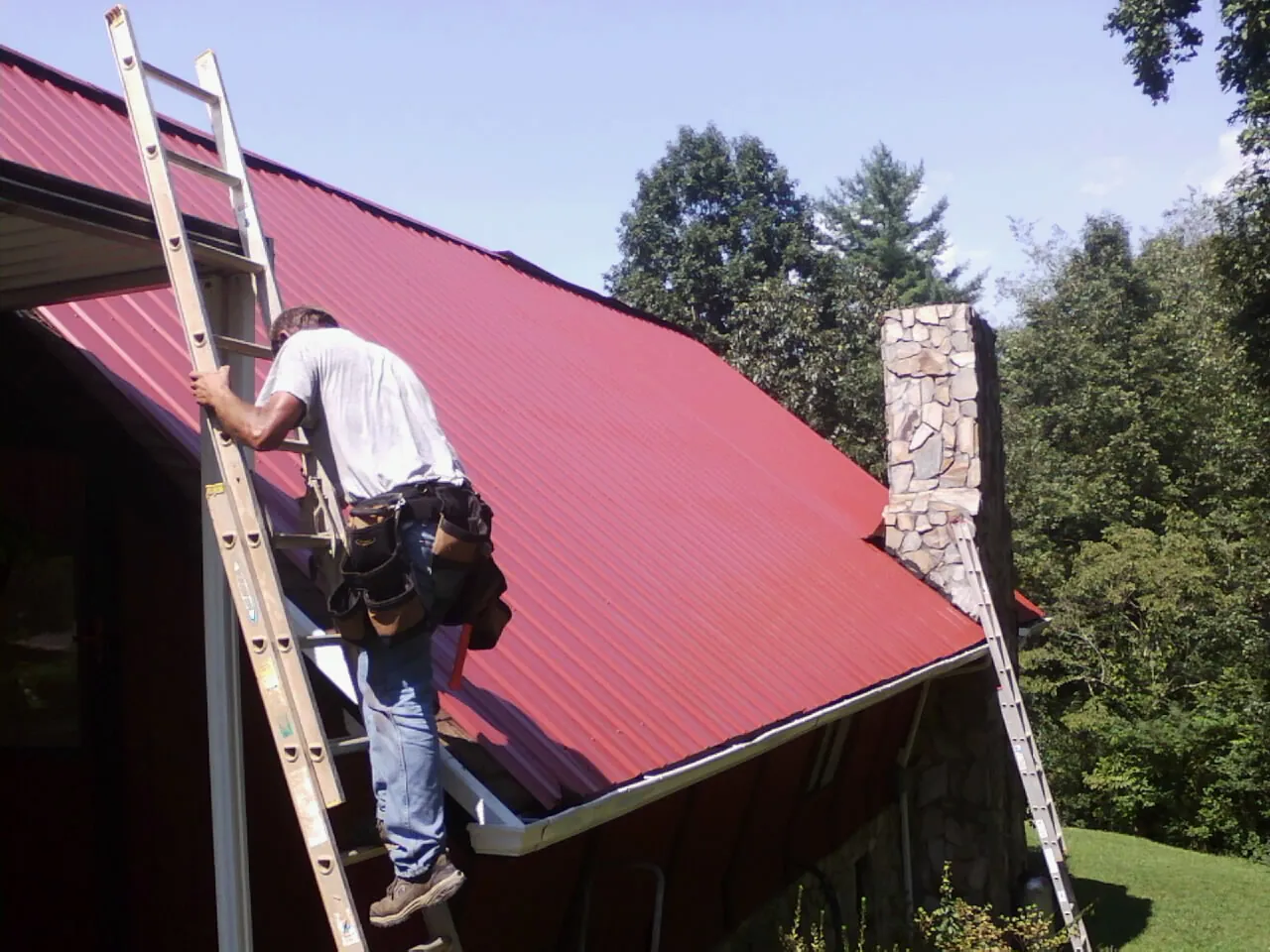 Roofer on a ladder beside a red metal roof and stone chimney — Soffit & Fascia Repair by Roofing Brothers in Lyndora